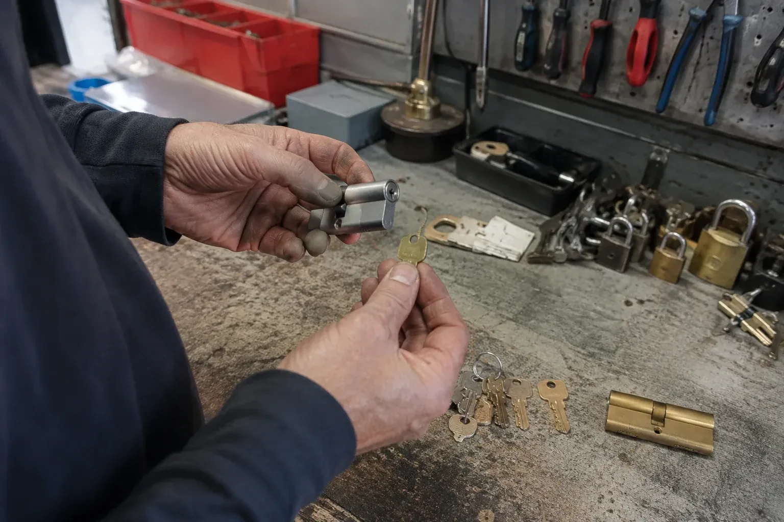 Hands holding a silver-colored lock cylinder and a brass-colored key above a workbench with padlocks, keys and tools. — Master Key System Design, Newmarket